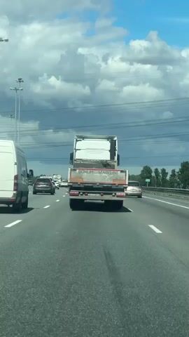 This is a first-person perspective video clip shot from a vehicle driving on a highway. The scene is set on a multi-lane road with a white van in the left lane and a white flatbed truck in the right lane, with other cars also visible. The truck has the words "BONUM MAIN LINE" printed on the back. The weather appears to be cloudy with some patches of blue sky. The time of day is likely afternoon, given the lighting. The overall mood of the video is mundane, depicting typical highway traffic. The location is identified as Zanevka, Russia.