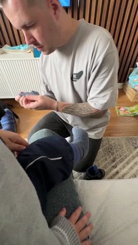 A man is shown in a medium shot, kneeling and putting socks on a child who is sitting on another person's lap. The man is wearing a light grey t-shirt, black pants, and a silver bracelet. He has a tattoo on his left forearm. The child is wearing blue socks, blue pants with a white stripe, and a blue top. The person whose lap the child is sitting on is wearing a grey sweater. The background includes a white cabinet, a wooden wall, a rug, and children's shoes. The camera is held at chest level, providing a close view of the interaction.