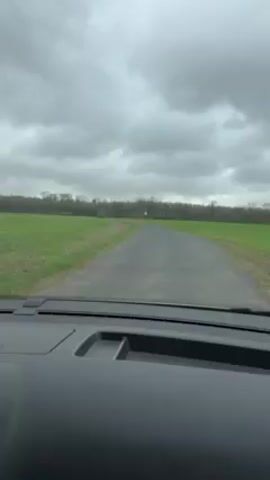 The video shows a view from inside a vehicle driving down a narrow asphalt road. On either side of the road are flat, green fields. In the distance, a line of trees can be seen. The sky is overcast and gray. The dashboard of the vehicle is visible in the foreground.