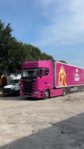 A bright pink Scania semi-truck and matching refrigerated trailer slowly drive from left to right across a paved outdoor area under a clear blue sky. The truck's cab features white trim and chrome accents. The pink trailer is adorned with Barbie branding, including a large graphic of Barbie's face with a crown at the front, followed by the text "Come on Barbie let's go party" surrounded by white party-themed graphics. A smaller "Barbie" logo is visible near the rear of the trailer. Green trees line the background. A white delivery van is partially visible behind the moving trailer.