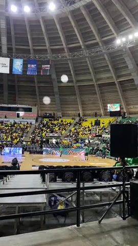 The video shows an indoor basketball game in progress. The camera is positioned in the stands, looking down at the court. The stands are filled with spectators wearing predominantly yellow and green shirts. The court has "Gran Final" emblazoned on it in large, illuminated lettering. A player in a green uniform is dribbling the ball near the center of the court, preparing to make a shot. Another player in a similar uniform is visible near the basket on the right side of the frame. The arena has a distinctive domed ceiling with visible wooden beams. Large banners hang from the ceiling, displaying logos and text that are not clearly legible. Speakers are visible in the foreground, suggesting the presence of a sound system for the event. The lighting appears to be artificial stadium lighting.