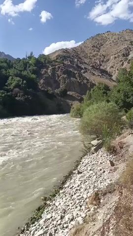 The video shows a wide, fast-moving river with muddy water flowing through a mountainous area. The riverbanks are rocky and covered with sparse vegetation. Steep hillsides covered with trees and shrubs rise on either side of the river. The sky is blue with some white clouds. The video appears to be filmed from a stationary position on the riverbank, looking downstream. There are no people visible in the video. The camera is handheld and the footage is somewhat shaky. The video is short and there is no discernible action other than the flow of the river.