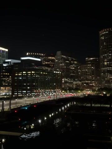 The video shows a nighttime cityscape with several tall buildings lit up against a dark sky. A bridge crosses a body of water in the foreground, reflecting lights from the city. Cars drive along the bridge. There is no one visible in the video. The scene has an urban, nighttime ambiance.