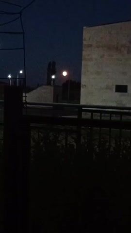 This is a video clip of a night scene in Aragatsavan, Armenia. The video opens with a view of a dark sky with a bright, full moon. In the foreground, there is a dark fence with some green bushes behind it. Further back, there are streetlights illuminating a paved area. There are also some buildings visible, one of which appears to be a light-colored concrete structure with dark windows. The overall mood of the video is quiet and peaceful, suggesting a calm evening. The time of day is clearly night, with the moon and streetlights being the primary sources of illumination. There is no visible text in the video.