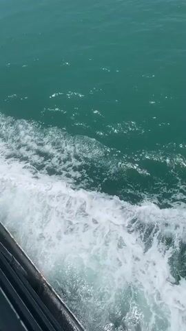 The video shows a view from the side of a boat moving through turquoise water. White foam churns along the side of the boat, indicating movement. Sunlight reflects off the water's surface. The boat itself is partially visible at the bottom left of the frame, with a dark railing. The water is clear enough to see faint shapes below the surface. The overall scene is bright and sunny.