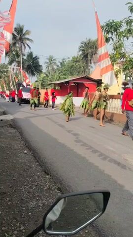 The video shows a procession on a paved road lined with houses and trees. Several people are walking in the procession. Some individuals are wearing red shirts, and others are dressed in costumes made of leaves and foliage, resembling natural outfits. Indonesian flags, red and white, are visible, decorating the area and adding a festive atmosphere. A white van is present in the background. The scene appears to be a cultural event or celebration taking place outdoors during the daytime, under a bright sky. The camera perspective seems to be from someone on a motorcycle, as a mirror of the motorcycle is visible in the foreground.