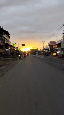 The video captures a street scene at dusk. The camera appears to be mounted on a moving vehicle, likely a motorcycle given the perspective and slight vibration. The road is asphalt and shows signs of wear. On the left side of the road, there are several buildings, including a multi-story structure and smaller shops, some with awnings. A few motorcycles are parked on the side and one is seen riding in the video. There is a street signpost with green directional signs. Power lines are visible overhead, strung between utility poles. On the right side of the road, there are more buildings, including shops and what appears to be a convenience store with an illuminated sign. Several cars and motorcycles are visible on the road, some moving and some parked. The sky is overcast with a dramatic sunset creating a warm orange and yellow glow on the horizon. The overall impression is a busy street in a town or city during the transition from day to night.