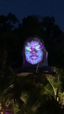 At night, a statue of a person with facial mapping is visible in the darkness, above the treeline. The statue is partially obscured by trees in the foreground, mostly palm trees. The facial mapping consists of bright neon colored patterns and lines in shades of green, purple, blue and white. The mapping is focused on the eyes, nose and lips. A blue light illuminates the statue.