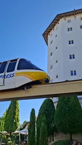 A monorail train, labeled "E.P. EXPRESS" in blue lettering with yellow stripes, travels along an elevated track against a clear blue sky. A building with a light-colored facade and brown trim is visible on the right side of the frame, featuring multiple windows. Below the monorail track, lush green trees and some foliage are present, with a paved area and a dark blue car in the lower portion of the image. The scene appears to be outdoors on a sunny day.