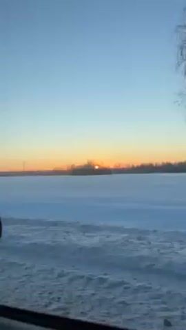 The video shows a snowy landscape during a sunset, as seen from the window of a moving car. The sky is a gradient of blue to orange, with the sun appearing as a bright spot on the horizon, partially obscured by trees. A snowy field stretches out to the horizon. The road next to the car is also covered in snow.  A side view mirror is visible. Trees with bare branches are visible in the foreground.