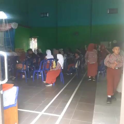 The video shows an indoor scene, likely a hall or auditorium. The walls are painted green, and the ceiling is turquoise. Rows of blue plastic chairs are arranged on a dark tiled floor, with white tape marking lanes or boundaries on the floor. Several people are seated in the chairs, mostly women and girls wearing hijabs and long dresses or skirts. Some are facing forward, as if watching something, while others appear to be interacting amongst themselves. On the right side of the frame, a young boy in a patterned shirt and red pants stands facing away from the camera, seemingly observing the scene. The lighting appears to be natural and artificial, suggesting it is daytime. There is a slight blur and low resolution to the video, making it difficult to discern specific details about faces or objects. The overall atmosphere suggests a gathering or event, possibly a school function or a religious ceremony.