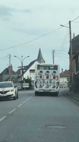 The video shows a view from a vehicle driving on a paved road in a village. A white bus is in front of the vehicle, traveling in the same direction. The bus has a license plate and a visible destination sign displaying "710". On the left side of the road, there is a white building with a blue roof and a steeple. The building has signs that read "A LOUER" (for rent) and "VLT Auto Mecanique Automobile". Several cars are parked in front of this building, including a black SUV and a silver hatchback. There are also street signs indicating directions to various towns. Utility poles with numerous wires are visible along the roadside, and the sky is overcast. The overall scene depicts a typical rural or suburban environment with everyday traffic and buildings.