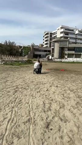 A man pushes a baby stroller on a sandy area, likely a beach, towards the camera. He is wearing a green t-shirt, dark pants, and sunglasses. The stroller is white with a dark frame. In the background is a multi-story building with a sign that reads "Marossa." Some trees and a stone wall are visible to the left, with other people and bicycles near the building in the background. The sky is cloudy. Tire tracks are visible in the sand.