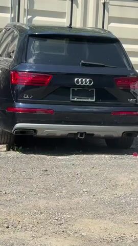 A dark blue Audi Q7 SUV is parked on a gravel surface in front of a white corrugated metal structure. The rear of the vehicle is visible, including the taillights, the Audi logo, and the "Q7" badging. A trailer hitch receiver is also visible on the rear bumper. The lighting suggests it is daytime.