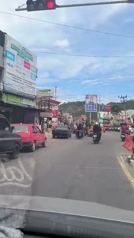 The video captures a street scene in a town, likely in Asia, with a cloudy sky. Traffic is moderate, with several motorcycles, a few cars, and what appears to be a truck. People are seen riding on motorcycles and walking on the sidewalk. There are several buildings lining the street, including businesses with prominent signage, such as a computer store and a gas station. A traffic light is visible overhead, showing a red signal. The overall impression is a typical day in a bustling urban or semi-urban environment.