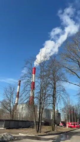 The video shows a daytime exterior shot of an industrial facility with two tall smokestacks emitting plumes of white smoke into a clear blue sky. The smokestacks are painted with red and white stripes. In the foreground, there are bare trees without leaves, suggesting a cold season. A concrete wall runs along the front of the facility, and what appears to be a roadway is visible in the lower part of the frame. In the distance, a red fire engine and several flags on poles can be seen near what may be the entrance to the facility. The camera appears to be stationary, offering a wide view of the scene.