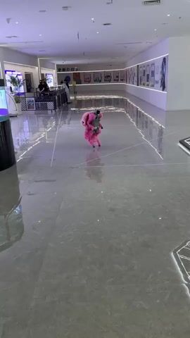 The video depicts a pink robot, resembling a small, furry creature, moving across a polished, gray-tiled floor. The robot has what appears to be a camera mounted on its back. It makes a sudden, jerky movement causing it to fall over. The robot is situated in a large, well-lit indoor space with a long corridor.  Along the corridor walls are displays containing images and text. People are seen walking in the background, seemingly unconcerned with the robot's actions. At the edge of the frame is a geometric installation on the floor.