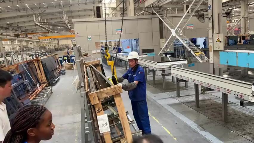 The scene is inside a well-lit factory, with a concrete floor and a high ceiling with visible beams and overhead cranes. Several people are present, including a worker in blue uniform, a white hardhat, and gloves, who is guiding a large pane of glass suspended by a chain connected to an overhead crane. The glass panel is being lifted from a wooden pallet. A woman in the foreground is taking a photo of the scene with her smartphone. Pallets loaded with similar glass panels line the left side of the frame. On the right side, there are long, white worktables with black tops, and some blue cabinets. Other workers can be seen in the background. Overall, the scene depicts glass production or assembly in a factory setting.