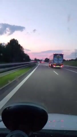 The video shows a first-person perspective of a drive on a highway at dusk. The sky is a mix of pink, purple, and blue, with clouds visible. Trees line the left side of the highway, and a guardrail separates the lanes. The highway is paved and has white lane markers. There are several vehicles on the road, including a large truck with an "Ice Tea" logo on its trailer, which is in the right lane ahead of the viewer. Other cars and trucks are visible further down the highway. The camera is positioned inside a vehicle, showing the dashboard and windshield. The overall atmosphere is calm and scenic.