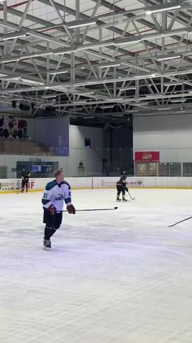 The video shows five hockey players in jerseys, with one in a gold jersey and four in turquoise jerseys, standing on an ice rink. They are gathered in a circle, seemingly talking or strategizing. The environment includes a large ice rink with visible lines and markings. Above them is a modern ceiling structure with numerous lights. In the background, there are advertisements and a scoreboard displaying the game details. There are some people skating by as well.