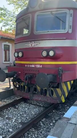 The video shows a maroon and silver electric locomotive parked at a train platform. The locomotive has the number "91 52 0044 150-8" painted on the front, along with a yellow stripe and a logo with the letters "SŽ."  A man wearing a dark jacket and holding a backpack walks along the platform next to the train. The platform is made of concrete slabs with blue and white painted edges.  The train tracks are visible next to the platform. In the background, there is a building with a window, as well as trees and a blue sky. The sun is shining.