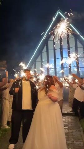 A wedding party is celebrating with sparklers at night in front of a building with a large, slanted window and a neon light outline. A bride in a white dress and a groom in a black jacket and white shirt are holding hands and smiling as guests surround them, holding up their own sparklers. The sparklers are emitting bright lights and sparks, creating a festive atmosphere. The building appears to be a modern structure, possibly a chapel or event venue. The ground is paved with bricks, and there is some greenery on the left side of the frame. The sky is dark, indicating it is nighttime. The overall scene is celebratory and joyous.