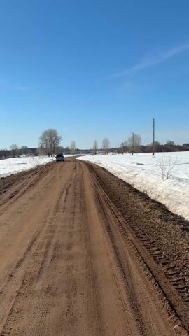 The video shows a dirt road in a rural area on a sunny day. The road is muddy and has tire tracks on it. On the right side of the road, there is snow and dry grass. On the left side of the road, there is a frozen body of water, with snow on its edges. In the distance, a car is parked on the road. A person is standing on the frozen lake in the middle of the video. The sky is blue with scattered clouds.
