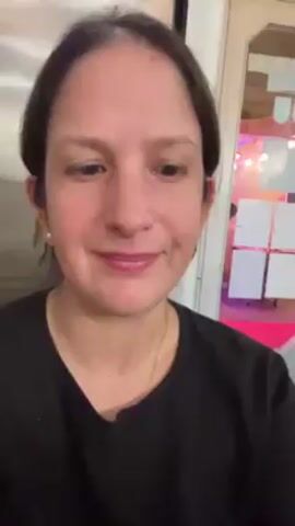 A fair-skinned woman with dark brown hair pulled back is centered in the frame from the chest up, smiling. She wears a black shirt and a gold necklace and has a small pearl earring. In the background is a door that opens to a view of pink and white decor. The woman gives a thumbs up and then makes a half-heart shape with her hand.