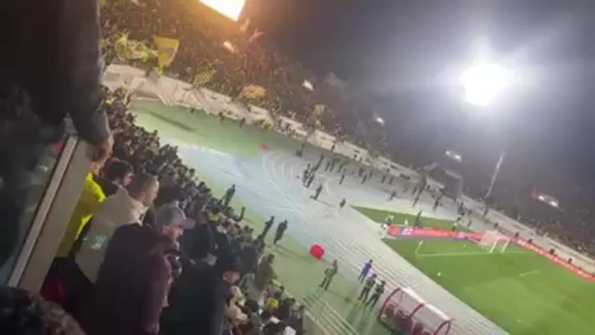 The video shows a football stadium at night, packed with spectators, many of whom are waving yellow flags. There are several people on the track encircling the football field, and a red object is visible near one of the goals. A dugout is situated along the sideline. The crowd is densely packed in the stands, and the atmosphere seems lively with a possible event or celebration taking place.