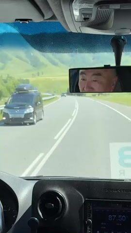 The video shows a view from inside a vehicle driving on a paved road. The dashboard is visible in the foreground. A rearview mirror reflects the face of a man with dark hair and eyes, wearing a dark jacket. The road curves to the right and is bordered by a metal guardrail. Beyond the guardrail are grassy hills with scattered trees. Another vehicle, a dark SUV, is driving in the opposite direction. The sky is overcast, and there are some green trees and bushes along the roadside. The landscape appears to be mountainous or hilly with lush green vegetation.