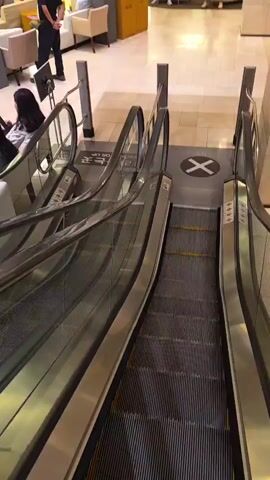 This is a high-angle video of two escalators going down side-by-side in a modern, indoor public space, possibly a hotel lobby or a library. The escalators are made of stainless steel with glass handrails. There are yellow safety lines on the escalator steps. On the floor between the escalators, there is a large black and white circular sign with a black "X" inside. In the background, several people are visible sitting on chairs and sofas in a lounge area. One person in a black shirt and black pants stands near the top of the left escalator, facing away from the camera. Another person in a black shirt and dark pants is sitting on a sofa in the lounge area, facing the camera. The overall environment appears clean and well-lit, with beige tiled floors and light-colored walls.