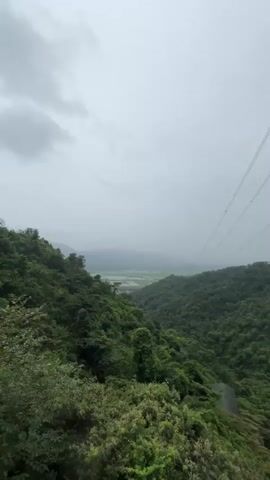 A drone flies over a valley with lush green trees. The sky is overcast. Several power lines stretch across the frame. A person in a black shirt and black pants stands on a balcony, looking out at the view.