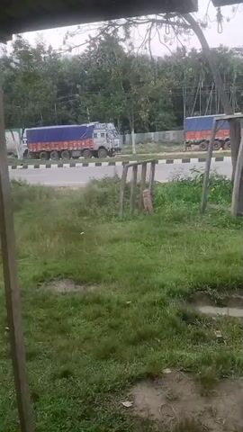 The video shows a grassy area in front of a road. Two large trucks are visible driving on the road, one in the foreground and one in the background. Both trucks are covered with blue tarps. The foreground truck appears to be a long, multi-axle vehicle. The background truck is partially obscured. There is a wooden structure in the foreground, possibly a swing set frame. The overall environment appears to be rural or semi-rural, with trees and vegetation present. The lighting suggests an overcast day.
