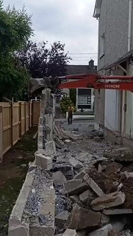 A demolition site with an excavator is shown. The excavator's arm is extended and its bucket is demolishing a concrete wall. Debris from the wall is scattered on the ground. A person in a yellow shirt is standing in the background. A wooden fence is visible to the left of the scene, and a house is visible to the right. The sky is overcast.