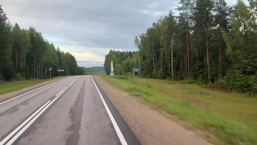 The video shows a road passing through a forest. On the right side of the road, there is a signpost with blue lettering that reads "КАДЫЙСКИЙ РАЙОН" (Kadyysky District) and a white obelisk with similar lettering. The road is paved and has white lines marking the lanes. The sky is cloudy. The video appears to be filmed from a moving vehicle.