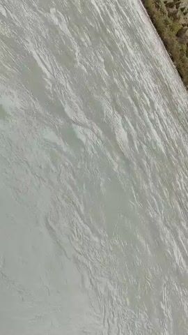 The video shows a wide, milky-gray river flowing through a mountainous valley under an overcast sky. The camera perspective is tilted, giving a sense of looking down at the water, which exhibits ripples and currents. On the right bank, there is a strip of dry, sparse vegetation and what appears to be a mountain slope. On the left bank, there is a patch of green grass and a rocky shoreline. Further in the distance, large, rugged mountains rise, some with snow-capped peaks visible through the clouds. The overall impression is a remote, natural landscape dominated by the river and mountains.