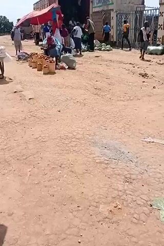 The video shows an outdoor market scene. A person is sitting on a chair in front of a display of buckets filled with potatoes. There are several other people around, some standing and some sitting. Various goods are being sold, including produce like watermelons, and packaged items. The ground is dirt and uneven. There are several umbrellas providing shade for the vendors. A cart with white bags is also visible. The scene appears to be a busy marketplace.