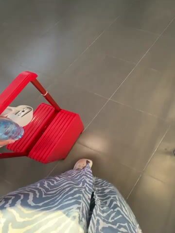 The video shows a first-person perspective of someone standing on a grey tiled floor. To the left, there is a red foldable cart with a bottle of water on it. A person's legs are visible wearing a blue and white zebra-patterned garment and white slippers. Further in the background, another person is walking away, carrying a straw bag filled with white items. The environment appears to be indoors, possibly a hallway or waiting area, with metal railings and a doorway visible. The camera pans slightly as the person holding the cart moves.