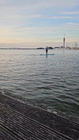A person is paddleboarding on a body of water. They are standing on a paddleboard and using a paddle to move through the water. The person is wearing a dark-colored shirt and shorts. In the background, there is a tall tower and several buildings. The sky is overcast with some clouds. The water appears calm with small ripples. In the foreground, there is a wooden dock or pier. The overall scene suggests a recreational activity in an urban or coastal environment.