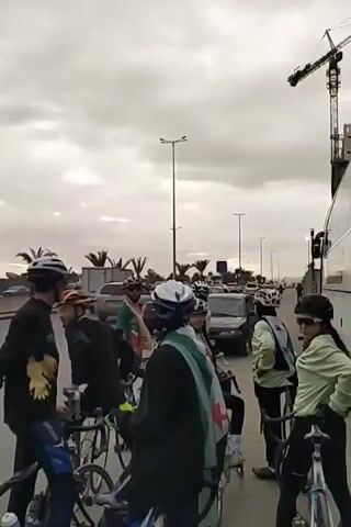 The video shows a group of cyclists standing on the side of a road, likely preparing for a ride or a rally. They are all wearing helmets and cycling gear. Some of them are holding up flags, which appear to be Palestinian flags. The cyclists are on a road next to city street with passing cars. The sky is overcast. The camera follows one cyclist, who is wearing a helmet, mask, and Palestinian flag.