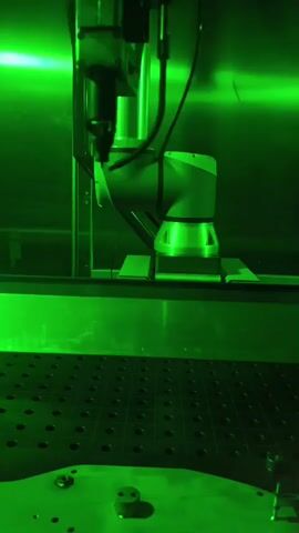A close-up, low-angle shot shows a robotic arm in a green-lit industrial setting. The arm, composed of metallic segments and cables, is positioned over a flat, perforated surface. The arm's end effector, a complex tool with a nozzle-like component, is directed downwards, almost touching the surface. In the background, another part of the robotic arm or machinery is visible. The overall impression is one of automated precision within a controlled environment, possibly involved in manufacturing or assembly.