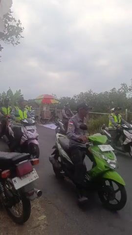 A group of police officers on motorcycles are riding down a paved road. The officers are wearing high-visibility vests and police uniforms. Some of the motorcycles are pink, green, and white. There are trees and vegetation on the side of the road, and the sky is cloudy. There is a white sheet or tarp on the ground behind some of the motorcycles. One officer appears to be speaking to another. The overall scene suggests a police escort or a response to an incident.