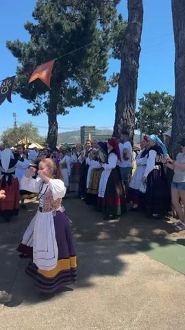 This is a vertical, outdoor, daytime video of a group of people, mostly young girls, dancing in a circle. The dancers are wearing traditional folk costumes consisting of long skirts, blouses, and head coverings. The skirts are layered and brightly colored, with prominent ruffles and embroidery. The blouses are white with puffed sleeves. Some of the girls have their hair in braids. They are dancing in what appears to be a village square or outdoor event space. The ground is paved with a light-colored, rough surface. In the background, there are trees, tents, and a crowd of spectators. There are also colorful flags or banners strung between trees and poles, adding to the festive atmosphere. The sun is shining brightly, casting shadows on the ground. The overall scene suggests a cultural festival or folk dance performance.