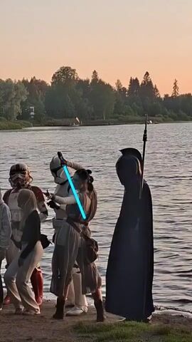 This video shows a group of people gathered near a body of water, possibly a lake or river. The background consists of trees and a clear sky with the sun setting, indicated by the warm lighting. Several individuals are dressed in costumes from the Star Wars franchise. Specifically, there are at least two individuals in Stormtrooper armor and one person dressed as Rey from Star Wars, holding a blue lightsaber. Another person is dressed in dark robes, resembling a Sith Lord, and holds a spear-like weapon. Some onlookers are not in costume. The costumed individuals appear to be interacting, with Rey and a Stormtrooper engaged in what looks like a mock lightsaber duel. The overall scene suggests a fan gathering or event.