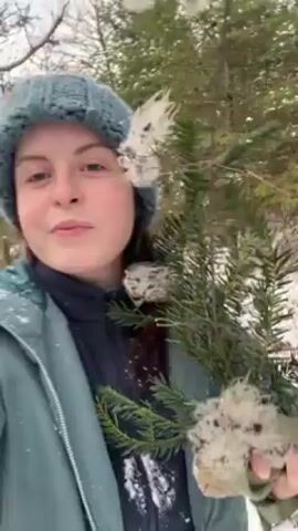 A woman with fair skin and green eyes, wearing a blue knitted hat and a green jacket over a dark turtleneck, holds evergreen boughs covered with white fluff in a snowy woodland setting. The camera is close to her face as she looks directly at it with a slight smile. Snow covers the ground and branches are visible in the background. The video is taken in daylight, with diffused light falling on the scene.