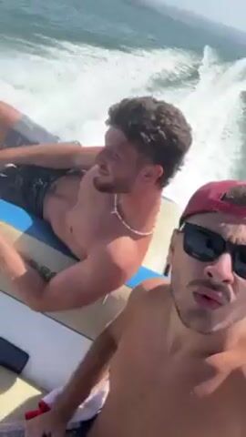 Okay, here's a detailed description of the video:

**Scene:** A sunny day on a motorboat moving quickly across the water. The camera appears to be held by someone on the boat.

**People:** Two men are visible in the frame. 
    * **Man 1 (Camera Holder):** Is in the foreground. He is shirtless, wearing black sunglasses and a red baseball cap. He has short dark hair and facial hair. He is looking directly into the camera, making facial expressions.
    * **Man 2:** Is in the background. He is shirtless, wearing grey patterned shorts and a white beaded necklace. He has curly brown hair and is lying down on the boat. He is looking off to the side, seemingly enjoying the ride.

**Objects:** 
    * **Motorboat:**  The men are on a motorboat. The boat is creating a wake.
    * **Water:** The boat is traveling on blue water.
    * **Sunglasses:** The man in the foreground is wearing sunglasses.
    * **Baseball Cap:** The man in the foreground is wearing a baseball cap.
    * **Necklace:** The man in the background is wearing a beaded necklace.
    * **Clothes:** Both men are wearing shorts.

**Actions:**
    * **Boating:** The boat is moving quickly across the water.
    * **Filming:** The man in the foreground is holding the camera and filming a selfie-style video.
    * **Relaxing:** The man in the background appears to be relaxing and enjoying the boat ride.

**Interactions:**
    * The two men are together on the boat. There is no direct interaction between them, but they are clearly sharing the experience.

**Environment:** The weather is sunny. The water is blue. Land is visible in the distance.
