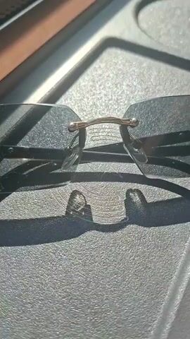 This is a close-up, overhead shot of a pair of frameless eyeglasses resting on a dark, textured surface. Sunlight casts shadows of the glasses and a thin strand of hair onto the surface. The eyeglasses appear to be made of clear lenses and a thin metal bridge. The background is out of focus but suggests an outdoor setting with a brown object in the upper left corner. The video is short and lacks any discernible action or interaction.