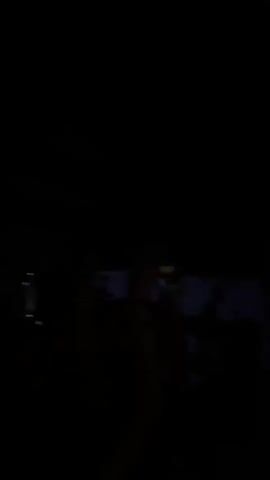 The video shows a dark, dimly lit scene, likely a concert or club, with a crowd of people. The lighting is mostly purple and blue, with some yellow light in the foreground. Several individuals are visible, with their arms raised and gesturing. One person in the foreground, lit by yellow light, appears to be singing or shouting. Another person in the middle ground, illuminated by blue light, is making a hand gesture that resembles the "rock on" sign. The background is dark, but some shapes that appear to be on a wall or screen are vaguely discernible, possibly illuminated by the stage lights. The overall atmosphere seems energetic and celebratory.