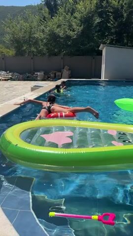 A person in a black swimsuit is lying on their stomach on a red inflatable raft in a swimming pool. Their arms are extended out to the sides, touching the edge of the pool. Their legs are spread apart. In the foreground, a green inflatable raft is visible, partially submerged in the water. In the background, other people are in the pool, and trees and a white building are visible. The water in the pool is blue. The day is sunny.
