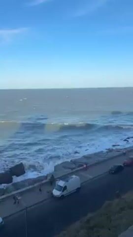 The video shows a coastal view under a clear, blue sky with sparse clouds. The ocean is active with waves crashing against a rocky coastline. A road runs parallel to the coast, featuring a sidewalk with a few people walking on it. Several vehicles are parked along the road, including a white van and at least two cars. The camera angle is elevated, providing a wide view of the seascape and the adjacent street.